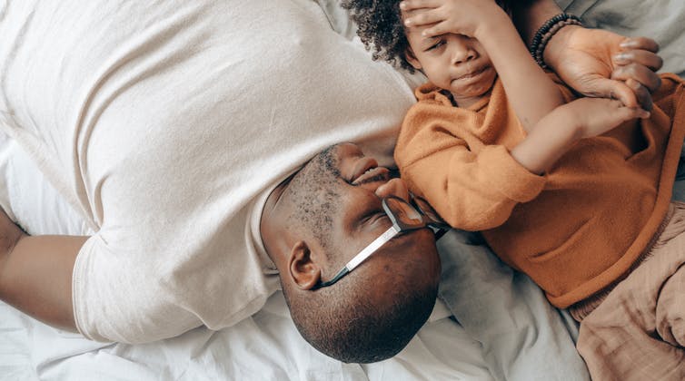 A father and child talk lying down and the boy looks a little apprehensive while the father holds his hand.