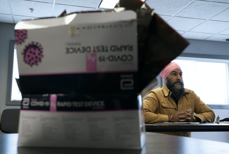 A COVID-19 rapid test package in the foreground, with NDP leader Jagmeet Singh seated at a table in the background