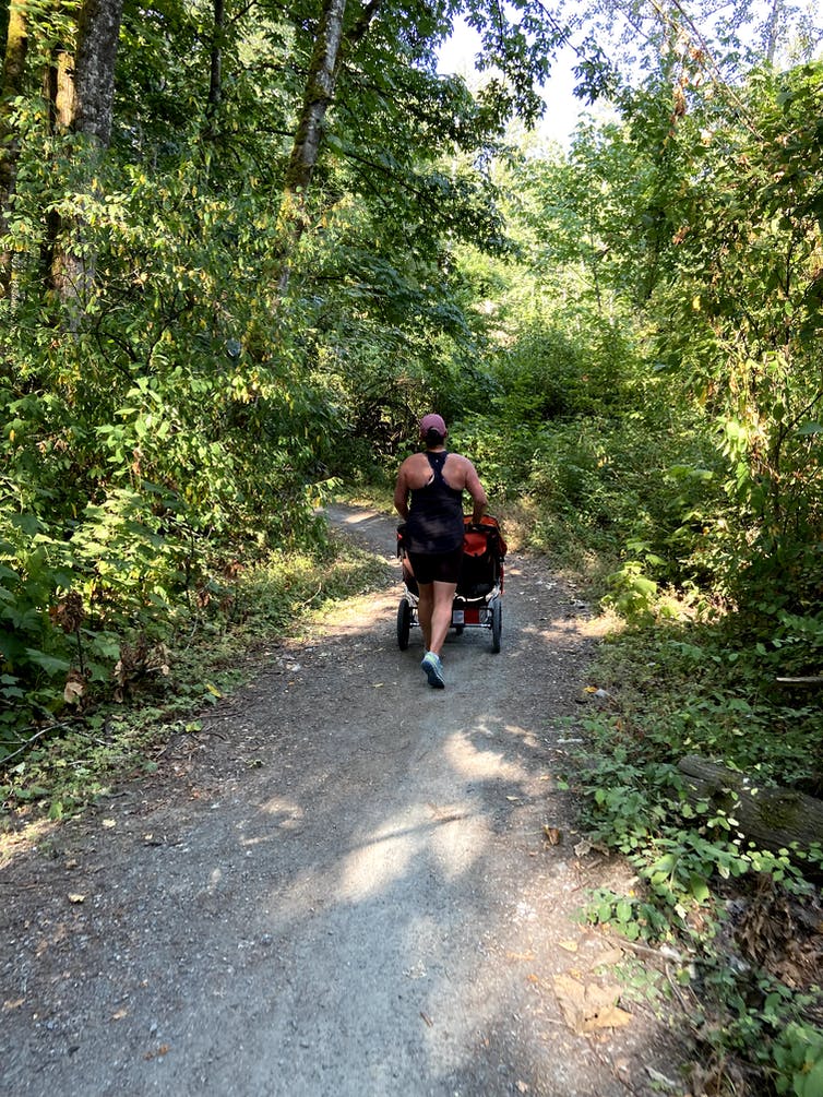 A woman seen from behind, jogging on a wooded path while pushing a double stroller