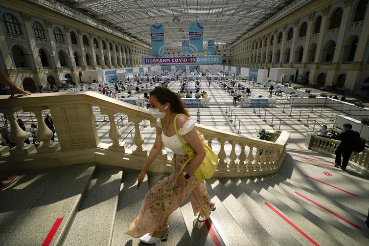 A woman climbs a curved staircase with an ornamental bannister. In the background is a vaccine clinic in a large open space with arches along the sides.