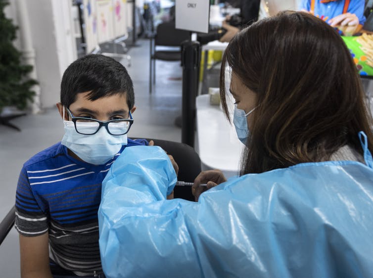 A boy wearing glasses and a mask gets vaccinated by a health-care worker wearing PPE