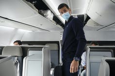A man with dark hair in a mask stands in an airplane.