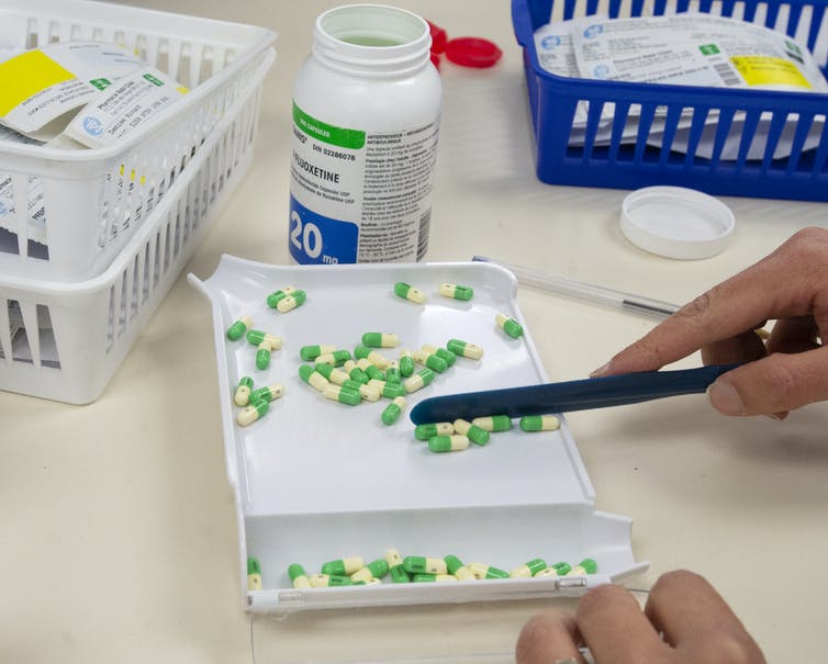 Pharmacist's hands using a pill tray to count green and yellow capsules, with a pill bottle and prescriptions in the background.