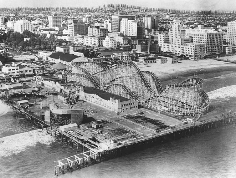 An old black-and-white photo of a roller coaster on a pier, with the city behind it and then a long row of oil derricks behind that on a ridge.