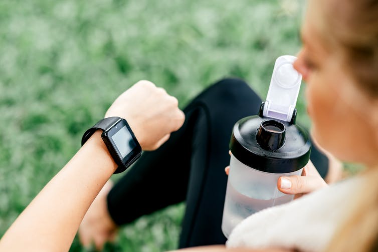 A woman holding a pre-workout shake in a plastic bottle looks at the smart watch on her wrist.