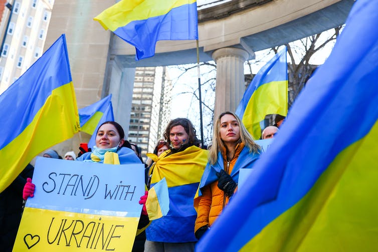 People waving Ukrainian flags and holding a sign painted like the Ukrainian flag reading 'Stand with Ukraine'
