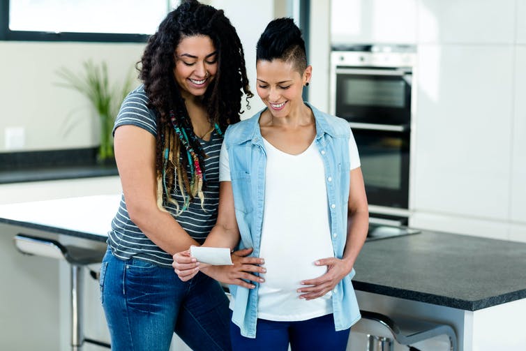 Two women standing in a kitchen looking at a small image, one is pregnant with her hand on her belly, the other has one the pregnant woman's belly and her other arm around her.