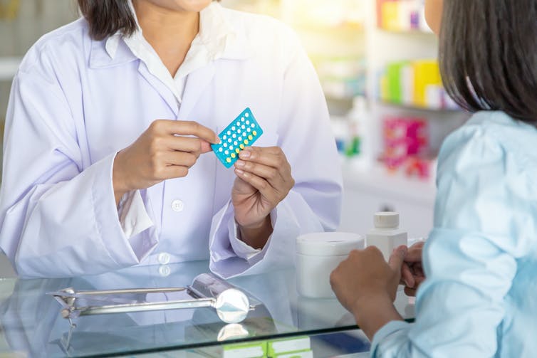 Cropped image of a pharmacist's hands holding birth control pills with a woman seen from behind
