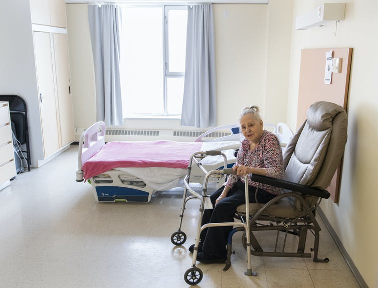 An elderly woman with her hair in a bun sits in a starkly furnished room in a long-term care facility, her walker in front of her.