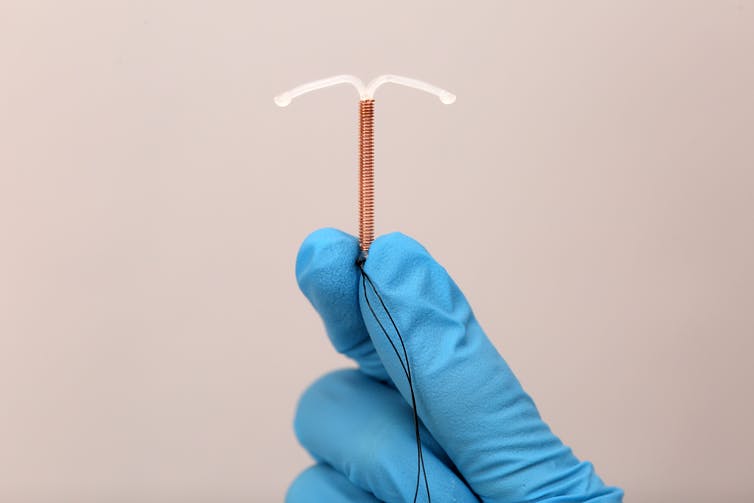A pair of hands wearing blue surgical gloves holds a copper IUD.