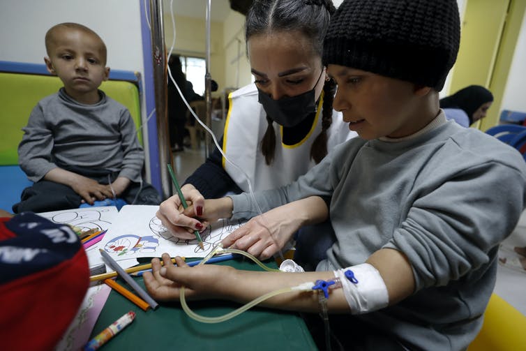 A boy in a hat with an IV line in his arm sits at a table drawing with a woman wearing a face mask as another child looks on