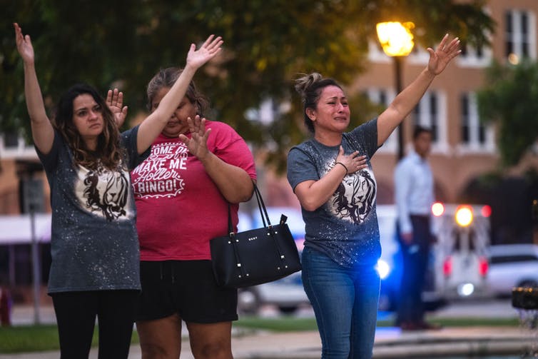 Three community members with grief-stricken faces hold their hands up toward the sky at a prayer vigil in Uvalde, Texas.