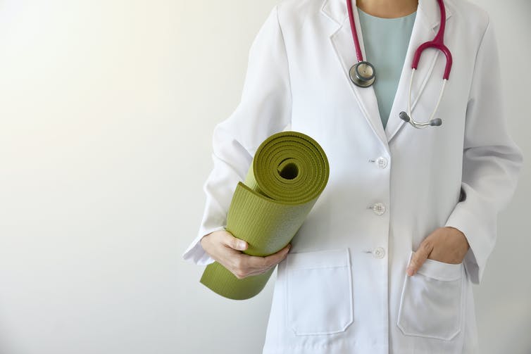 A person in a white coat with a stethoscope around their neck holding a rolled-up green yoga mat
