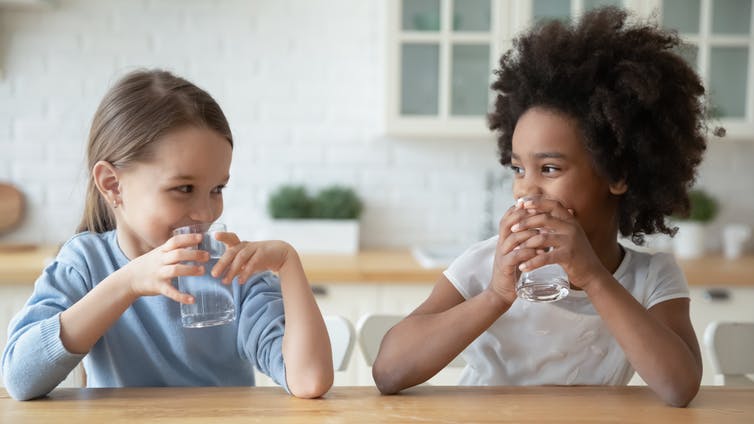 Two girls drinking water in a kitchen.
