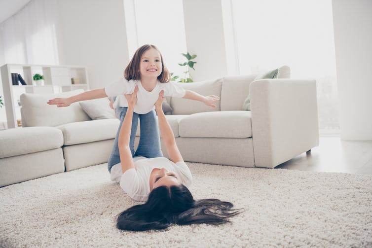 A woman lying on her back on white carpet holds up a little girl who is pretending to fly. A white couch is behind them.