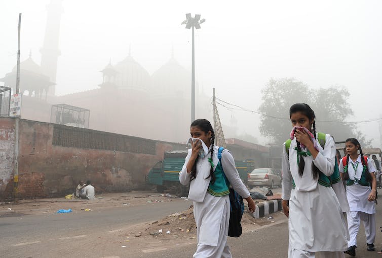 Three girls in white school uniforms walk down a smoggy street holding kerchiefs over their noses.