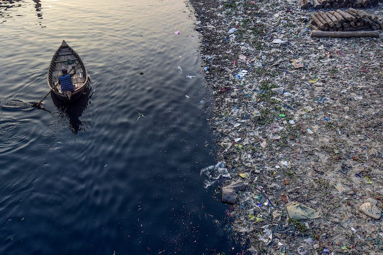 Viewed from above, a person paddles a wide canoe down a river lined with plastic and other trash.