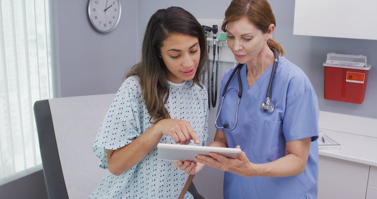 A woman in a hospital gown and a woman in scrubs and a stethoscope looking at tablet together
