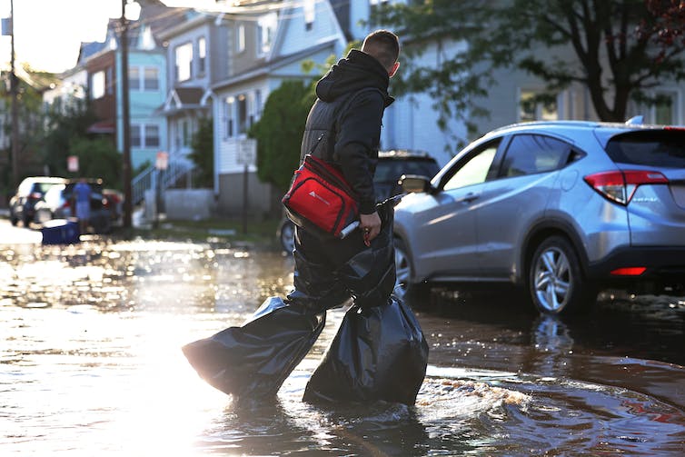 A person wearing trash bags around each leg to keep them dry crosses a flooded city street in New Jersey. An overturned trash can floats in the background.