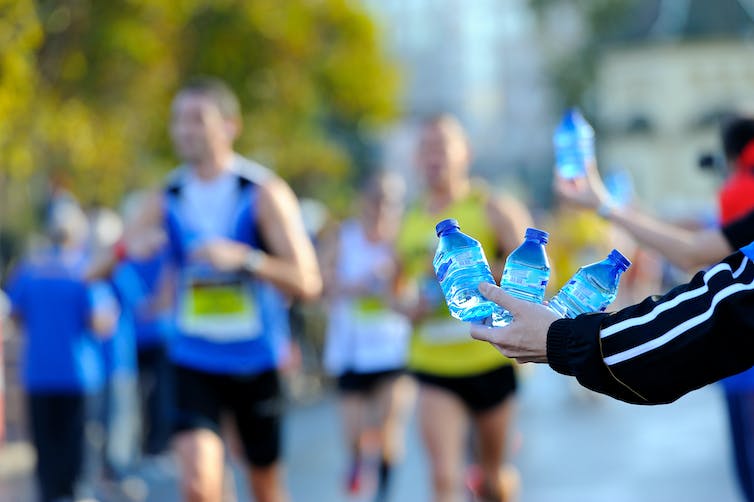 An arm offering three bottles of water to a group of runners who are out of focus in the background