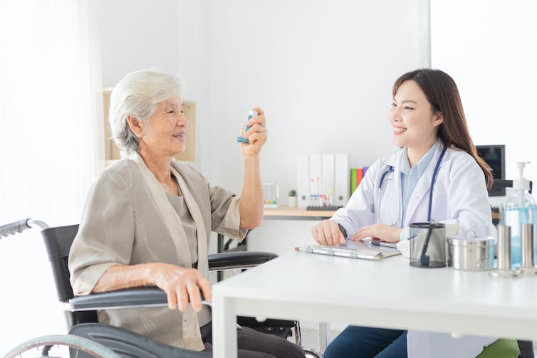 A young woman behind a desk in a white coat with stethoscope watching an older woman use an inhaler