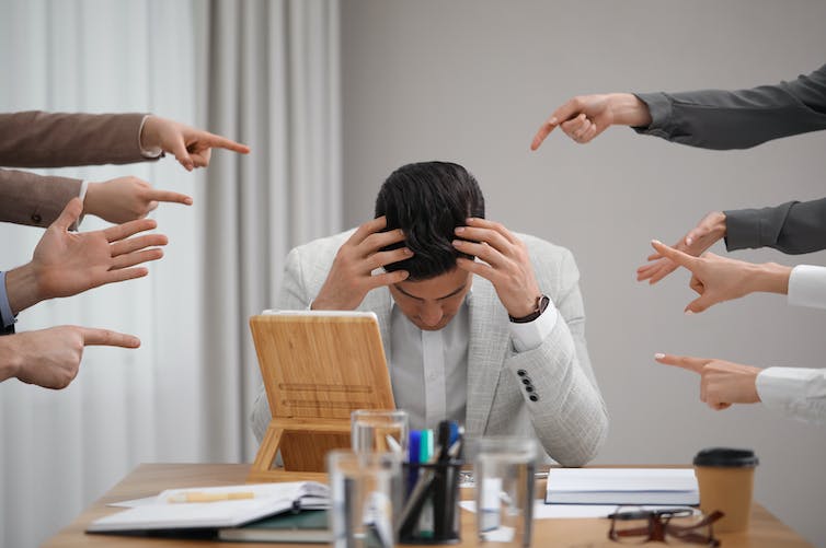 A man with his head down in his hands is surrounded by pointing fingers