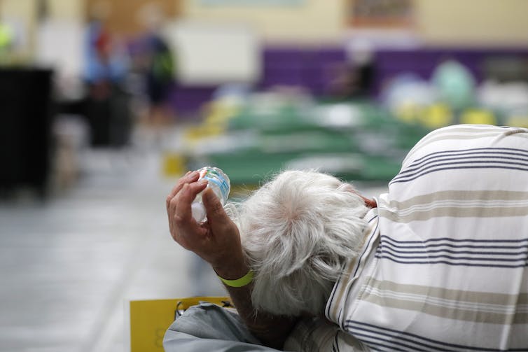 An older man lies on a cot in an evacuation center. He has a wrist band and is clutching a bottle of water.