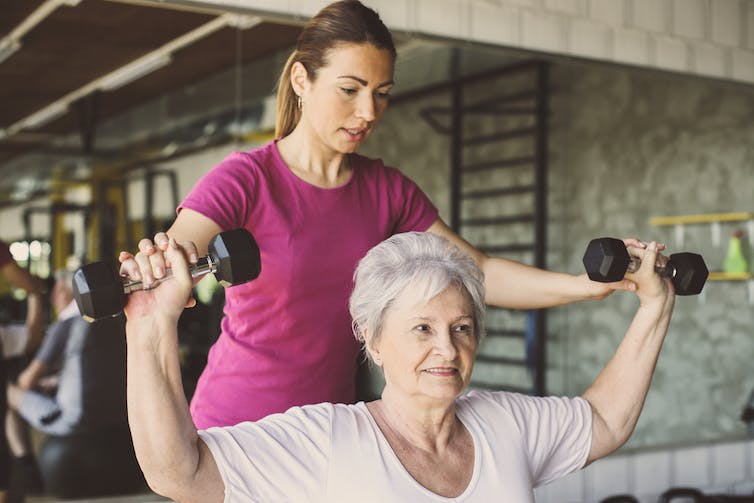 Older woman lifting dumbbells, assisted by a trainer