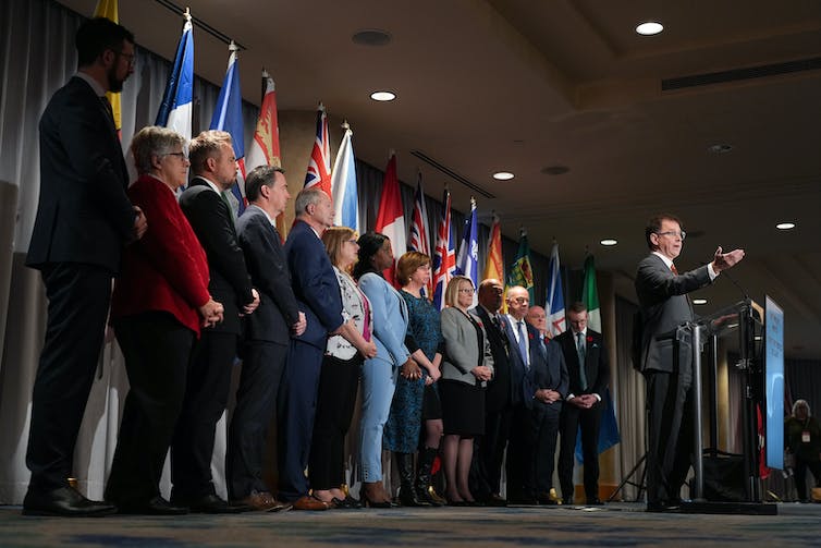 A man at a podium gesturing with his hand, and a line of people in business clothes behind him, with provincial flags