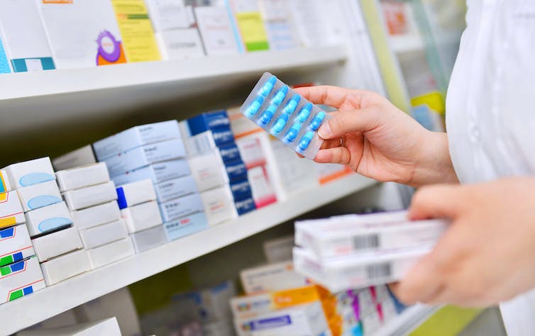 a shelf of packaged medications, the hands of someone wearing a white coat and holding a pill packet are visible