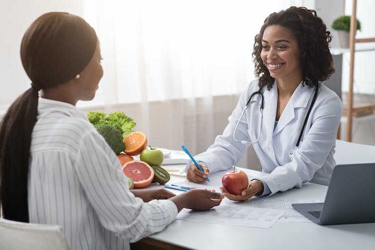A woman in a white coat holding an apple, with a bowl of fresh produce on her desk, consulting with a woman who has her back to the camera