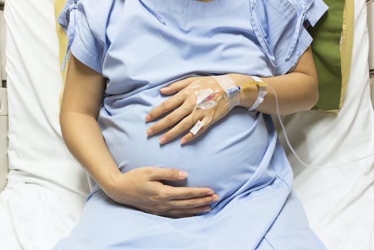 A pregnant woman in a hospital gown and bed holds her stomach.