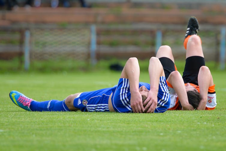 Two football players lie on the ground holding their heads.