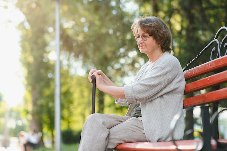A woman sits on a bench in a park holding a walking cane.
