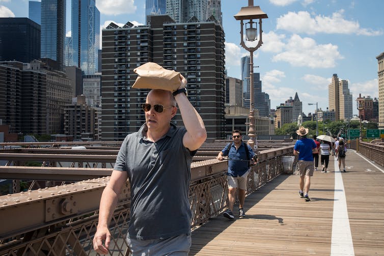A man holds something over his head to shield the sun from his forehead. Other people walking across the bridge on a bright, sunny day have umbrellas and hats.