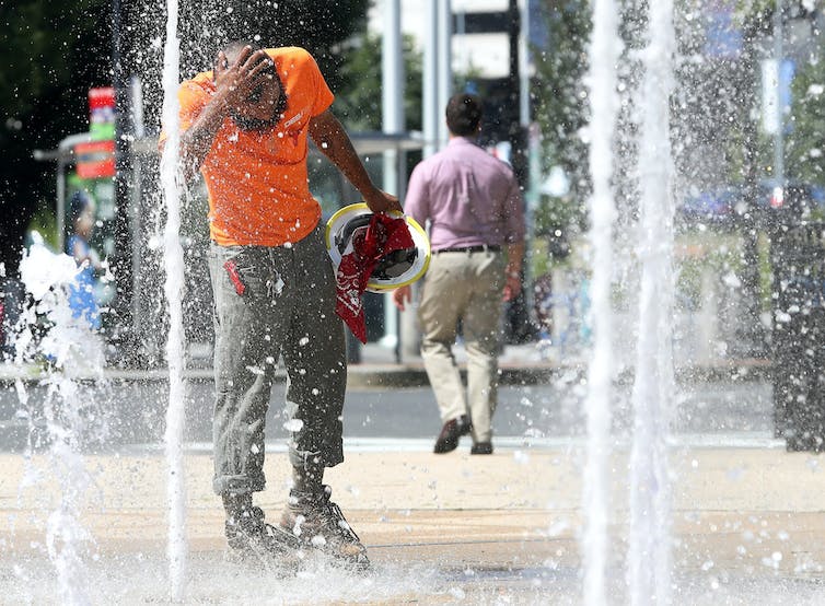 A construction worker cools his head in the stream of a fountain outside and office building.
