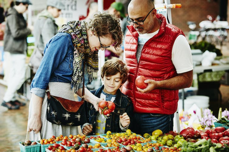 A male and female adult shop for vegetables with a child and examine tomatoes from a stall at a farmer's market.