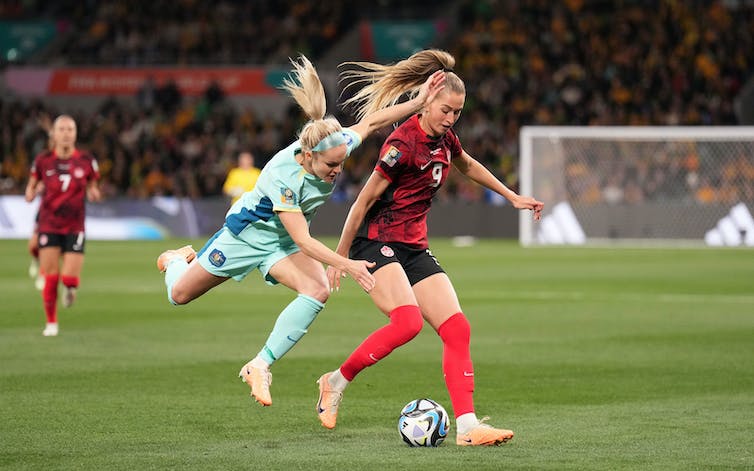 Two blonde soccer players fight for a ball, one wearing light blue and jumping into the left shoulder of one wearing red and black