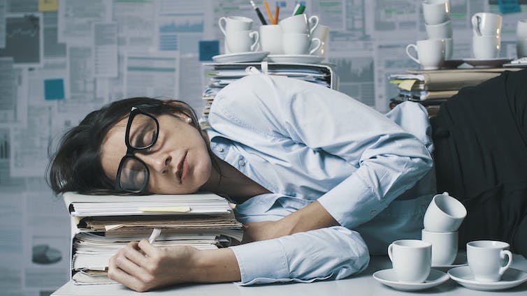Person with glasses sleeping on a stack of thick files, surrounded by coffee cups and paperwork