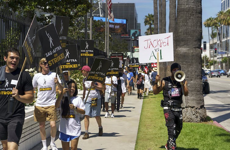 A line of people walk down a city sidewalk holding picket signs that say SAG-AFTRA ON STRIKE