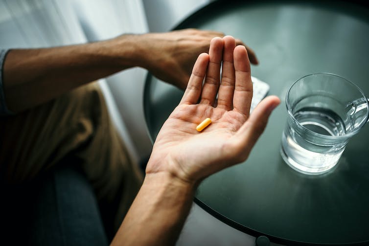 Close-up of hand holding pill beside a glass of water on a table