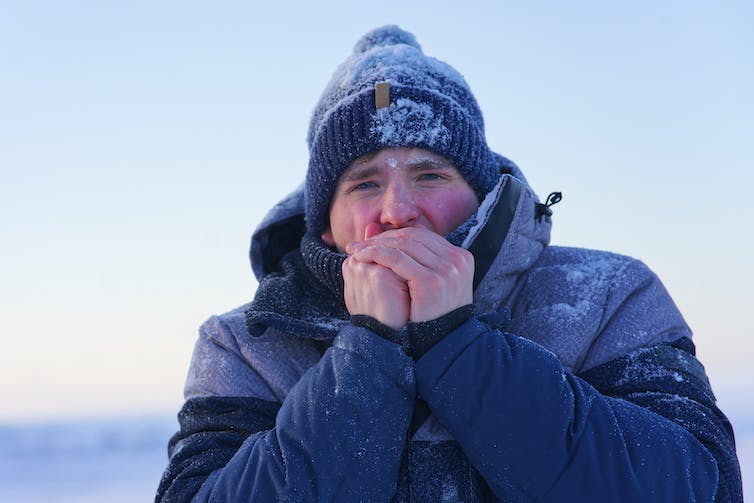 A man wearing a hat and winter jacket blows warm breath onto his cold hands while standing outside on a cold day.
