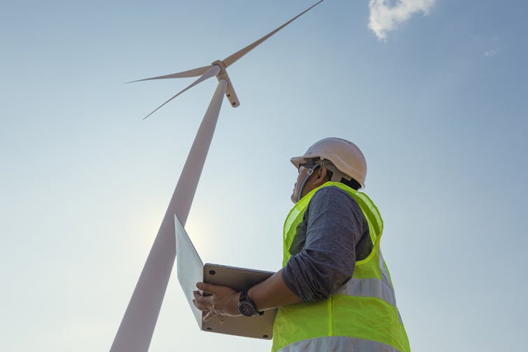 An engineer wearing a hard hat and bright green safety vest inspects a wind turbine.