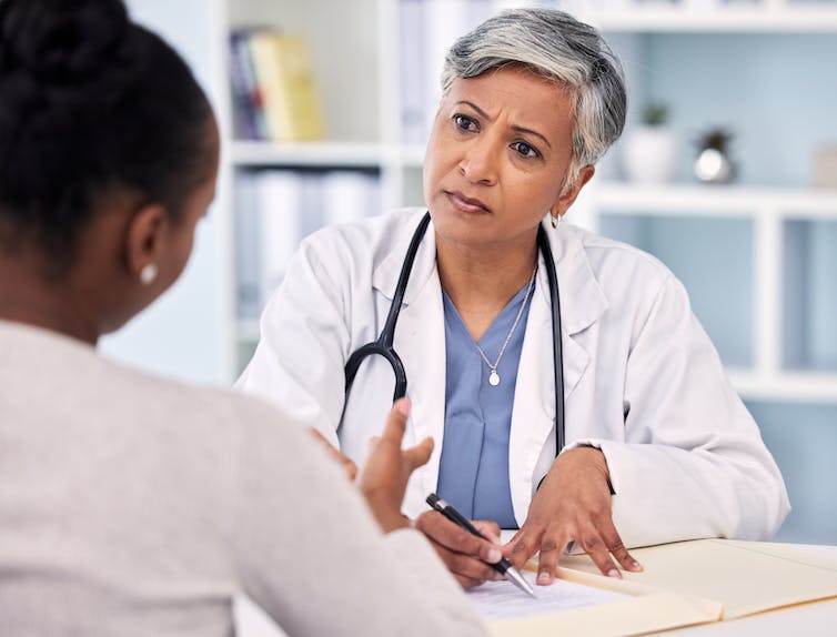 A woman with gray hair in a white coat and stethoscope listening to a person with their back to the camera