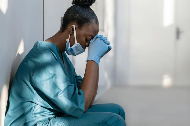 A stressed Black doctor in scrubs sits with her head resting on her hands.