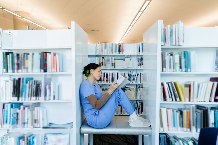 Clinician in scrubs sitting on a table between library shelves, reading a book