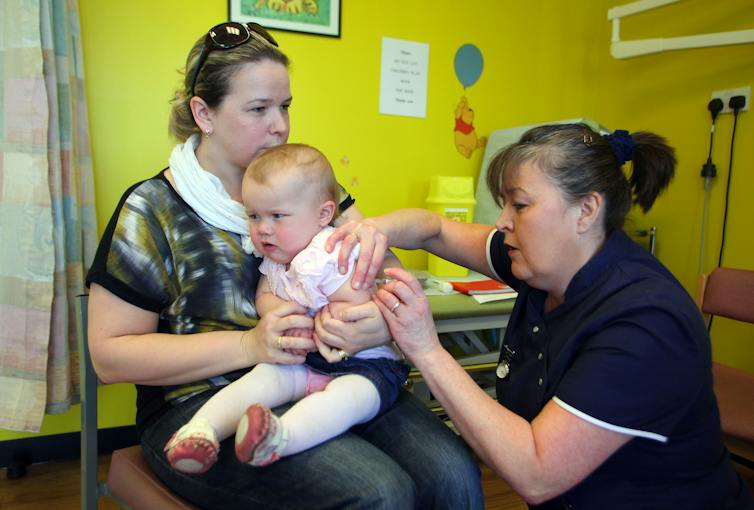Two women and baby are in a medical office with yellow walls. One of the women holds a baby, who's dressed in pink and looks unhappy, while the other woman, dressed in a nurse's uniform administers a vaccine to the baby.