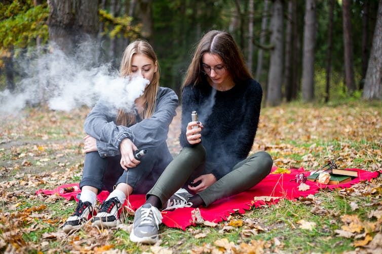 Two teen girls smoking electronic vapes and seated side by side on a picnic blanket in the woods