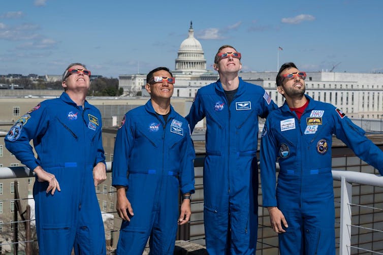 four astronauts in jumpsuits wearing eclipse glasses