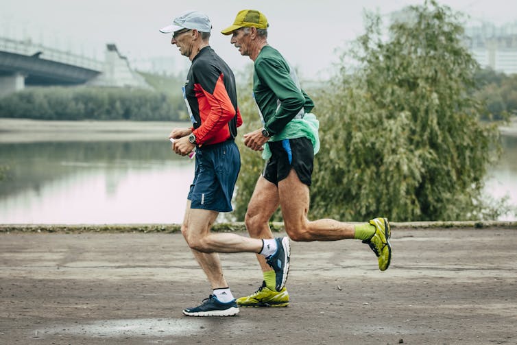 Two older men run along the side of a river.